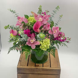 Pink calla lilies, orchids, and green hydrangeas in a clear glass vase on a wooden crate