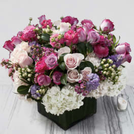 Low arrangement of pink and lavender roses with white hydrangeas in a green-wrapped glass vase.