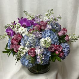 Lush arrangement of blue hydrangeas, pink roses, white stock, and purple orchids in a clear glass vase