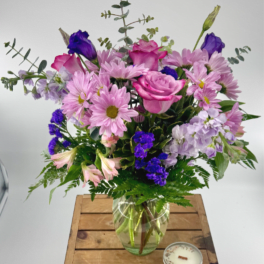 Mixed bouquet of pink roses and purple blooms in a glass vase on a wooden crate with a small candle.