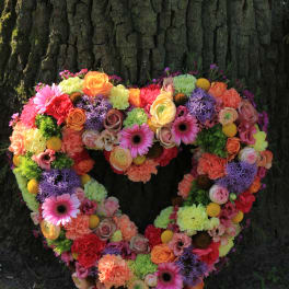 Heart-shaped wreath of bright mixed flowers in pink, orange, yellow, and purple leaning against a tree trunk