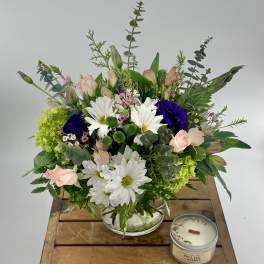 Mixed bouquet of white daisies, pink roses, and purple blooms in a glass bowl with a candle on a wooden crate