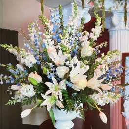 Tall arrangement of white lilies, roses, and blue delphinium in a white pedestal vase
