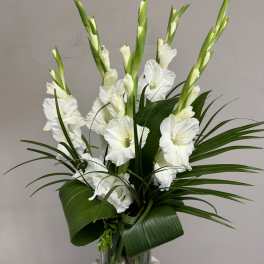 White gladiolus arrangement in a clear glass vase with broad green leaves