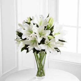 Tall bouquet of white lilies with green buds in a clear glass vase on a white table