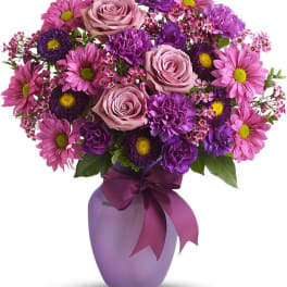 Purple and pink mixed flower arrangement in a lavender vase with a bow
