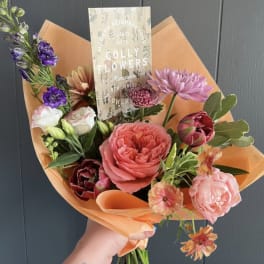 Hand-tied bouquet of pink, purple, and peach flowers wrapped in orange paper