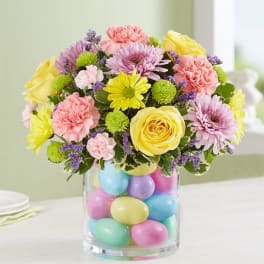 Pastel bouquet with yellow roses, pink carnations, and daisies in a glass vase filled with colorful eggs.