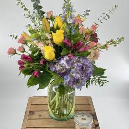 Tall mixed bouquet with yellow roses, lilies, hydrangea, and other flowers in a glass vase beside a small candle