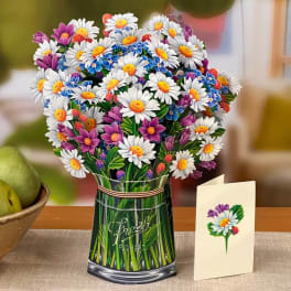 Vase of white daisies with purple and blue accent flowers beside a matching floral greeting card on a table