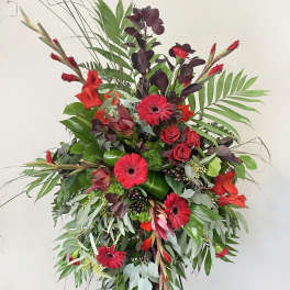 Tall standing spray of red gerbera daisies and roses with dramatic greenery on an easel.