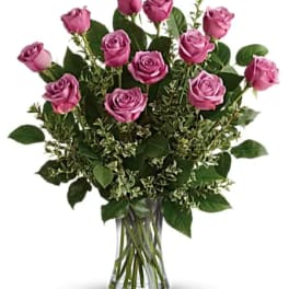 Dozen lavender roses in a clear glass vase with abundant filler foliage
