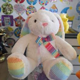 White plush bunny with rainbow ears, paws, and bow sitting in a gift shop display.