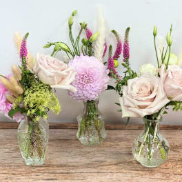 Trio of small glass bud vases with blush roses, a pink dahlia, and delicate accent flowers.