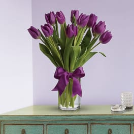 Tall bouquet of purple tulips in a clear glass vase with a matching purple ribbon, set on a green console table.