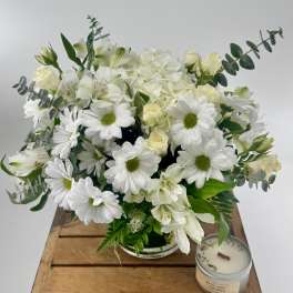 White daisies, roses, and hydrangeas in a glass vase beside a tin candle on a wooden crate.
