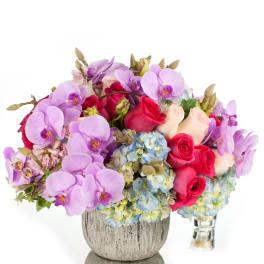 Compact arrangement of lavender orchids, pink and ivory roses, and blue hydrangeas in a gray vase