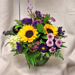 Bright arrangement with yellow sunflowers and mixed purple and pink flowers in a green bowl vase
