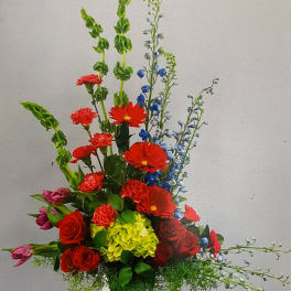 Tall mixed arrangement with red roses, gerbera, blue delphinium, and green hydrangea in a white vase