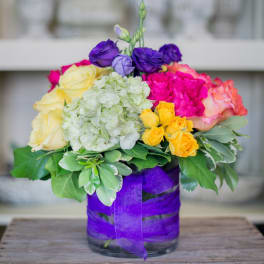 Colorful arrangement of purple lisianthus, yellow roses, and green hydrangea in a glass vase wrapped with purple ribbon
