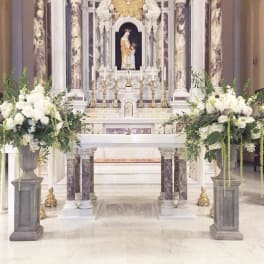 Large white floral altar arrangements flank a church altar with candles and a statue.