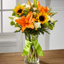 Bouquet of orange lilies, sunflowers, and white carnations in a glass vase with a green ribbon