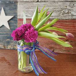 Tall glass vase with bright pink carnations and closed pink tulips, tied with a striped ribbon.