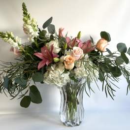 Pink lilies and roses in a clear glass vase with white blooms and greenery
