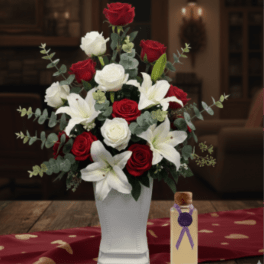 Tall arrangement of red and white roses with white lilies in a white vase beside a small gift bottle.