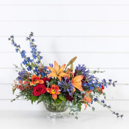 Mixed bouquet of orange lilies, red roses, and blue flowers in a glass vase