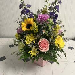 Mixed bouquet with pink rose, sunflower, purple blooms, and yellow daisies in a pink box