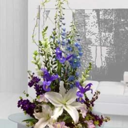 Tall arrangement with white lilies and purple-blue flowers in a low container