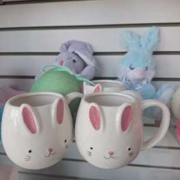 Two white bunny-face mugs with pink ears on a glass shelf with pastel bunny toys behind.