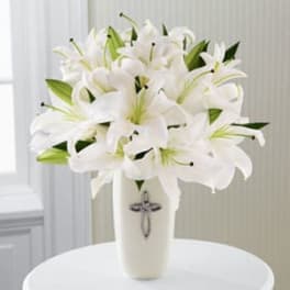 White lilies arranged in a tall white vase with a small cross emblem