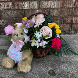 Teddy bear beside a basket of pink, yellow, and white flowers