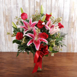 Tall arrangement of red roses and pink lilies in a clear glass vase with a red ribbon bow.