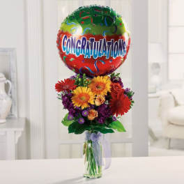 Tall mixed bouquet of bright gerbera daisies and roses in a glass vase with a Congratulations balloon.
