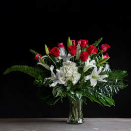 Tall arrangement of red roses, white lilies, and white hydrangea with arching green fronds in a clear vase.