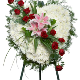 Heart-shaped standing floral spray of white flowers with red roses and pink lilies on a green easel.