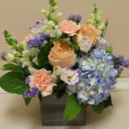 Mixed bouquet with blue hydrangea, peach roses, and purple accents in a square vase