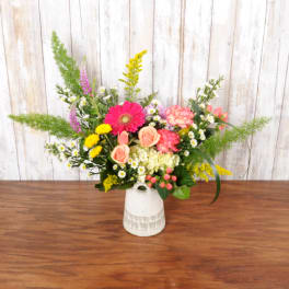 Colorful arrangement of pink gerbera, peach roses, and mixed blooms with tall greenery in a white ceramic jug.