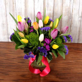 Low glass vase of mixed tulips with purple accent flowers and a pink ribbon bow.