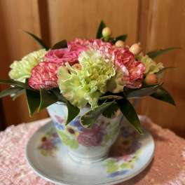 Small arrangement of pink roses and green carnations in a floral teacup with saucer on a lace cloth