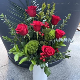 Red roses arranged in a white vase with greenery and red berries