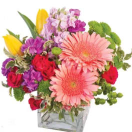Colorful mixed bouquet with pink gerbera daisies, red roses, purple blooms, and yellow tulips in a glass cube vase
