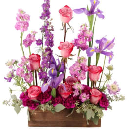 Pink roses, purple irises, and magenta blooms arranged upright in a rectangular wooden box.