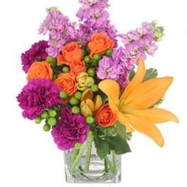 Bright arrangement of orange lilies and roses with purple flowers in a clear square vase