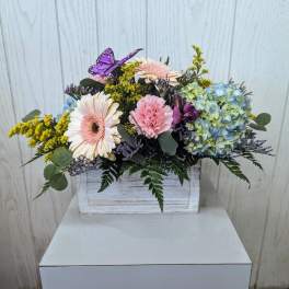 Mixed pastel flowers in a white wooden box with a purple butterfly accent.