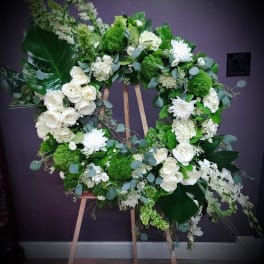 Large white and green standing wreath with roses, mums, orchids, and large glossy green leaves on a wooden easel.