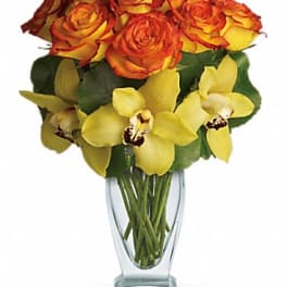 Tall clear vase with orange-yellow roses and yellow orchids in a compact arrangement